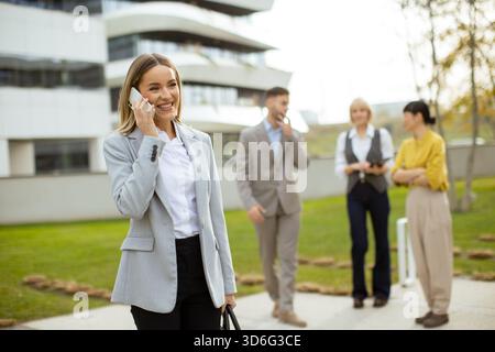Eine Frau lächelt und spricht am Telefon, während sie vor einem eleganten Bürogebäude steht. Im Hintergrund führen drei Kollegen Gespräche Stockfoto