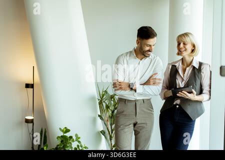 Zwei Kollegen verbringen einen leichten Moment in einem eleganten Büro. Sie lächeln und interagieren, während sie sich ein Tablet ansehen. Grüne Pflanzen verleihen dem W Stockfoto