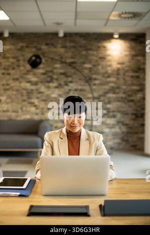 Eine Frau sitzt an einem Holztisch mit einem Laptop und konzentriert sich auf ihre Arbeit. Das moderne Büro ist mit Ziegelwänden und dezenter Beleuchtung ausgestattet und schafft eine komfortable Atmosphäre Stockfoto