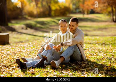 Koreanerin und kaukasischer Mann. Zwei Menschen teilen einen fröhlichen Moment, zusammen auf dem Gras in einem Park zu sitzen. Helle Herbstblätter sorgen für einen schönen Hintergrund Stockfoto