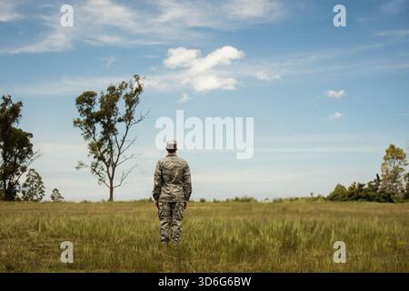 Männlicher Soldat, der mit Rücken zum Horizont auf dem Grasfeld steht, trägt Tarnuniform und Mütze. Militär, Grasland, Wiese, Himmel, Wolken, Stiefel, Hut Stockfoto