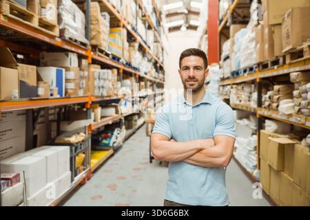 Mann in den späten 20ern mit hellblauem Polo, der im Lagerhaus zwischen Regalen und Kisten steht. Arbeiter, Logistik, Lagerung, Vertrieb, Industrie, PA Stockfoto