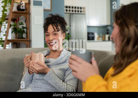 Verschiedene Freundinnen plaudern und trinken Kaffee auf dem grauen Sofa im Wohnbereich mit Keramikbecher Stockfoto