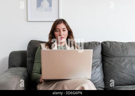 Frau, die auf silbernem Laptop arbeitet, sitzt zu Hause auf grauem Ledersofa mit abstrakten Kunstwerken Stockfoto