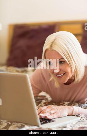 Reife Frau, die sich auf dem Bett liegend, mit einem hellrosa Oberteil, mit einem Ring, während sie einen silbernen Laptop benutzte. Schlafzimmer, Computer, blond, Kopfteil, Wärme, Entspannung Stockfoto