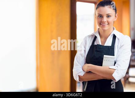 Weibliche Kellnerin steht im Café in weißem Hemd, schwarzer Schürze mit Bestellblock, Stift, Kopierraum. Gastfreundschaft, Gastronomie, Restaurant, Tageslicht, hell, fr Stockfoto