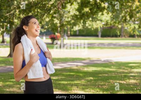 Frau in 20ern lacht, während sie im Park steht, Handtuch hält und Sportkleidung trägt, Kopierraum. Jogger, Outdoor, Natur, Sonnenschein, Pfad, offen, AT Stockfoto