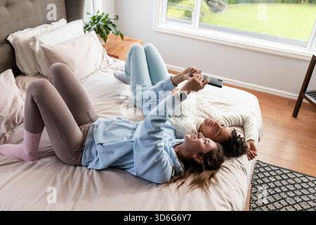 Verschiedene Freundinnen, die nebeneinander auf Bettdecken und Kissen liegen und das Smartphone am Fenster des Schlafzimmers halten Stockfoto