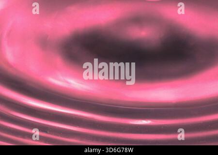259.Makrofoto von Wasserwelligkeit mit rosafarbenen Tönen und einem dunklen Mittelbereich, der durch einen Tropfenaufprall erzeugt wird, mit kreisförmigen Wellenmustern und glatten Reflexen Stockfoto
