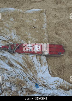Der Blick auf ein verwittertes rotes Rettungsschwimmer-Paddel, dessen Farbe abblättert und das Seil ausgefranst ist, ruht auf einem zerknitterten weißen Tuch vor der Kulisse von goldenem Sand, Punta Stockfoto