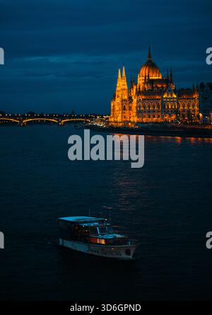 Budapest, Ungarn - 01. Februar 2025: Blick auf das beleuchtete ungarische Parlamentsgebäude auf der Donau, während ein Touristenboot durch die Donau gleitet Stockfoto