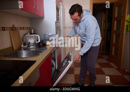 Ein Mann in einem leichten Kapuzenpullover lädt einen Geschirrspüler aus und gießt Kaffee in eine gemütliche Küche, um die tägliche Routine eines Vaters festzuhalten, der Arbeit und Familie in Einklang bringt. Stockfoto