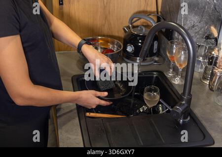 Person, die schwarze Schüssel in Küchenspüle wäscht. Eine Person wäscht eine schwarze Schüssel mit einem dunklen Schwamm unter fließendem Wasser an einer Küchenspüle, umgeben von Stockfoto