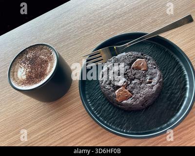 Heißer Cappuccino in einem schwarzen Keramikbecher serviert mit einem reichhaltigen Schokoladenkeks auf einem schwarzen Teller mit einer Gabel auf einem hölzernen Kaffeetisch in Malaysia Stockfoto