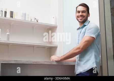 Erwachsener Mann, der sich auf eine rustikale Theke im Salon stützt, blaues Hemd trägt, in einem Regal mit Flaschen, Kopierraum. Holz, Display, Kosmetik, Boutique, minimal, br Stockfoto