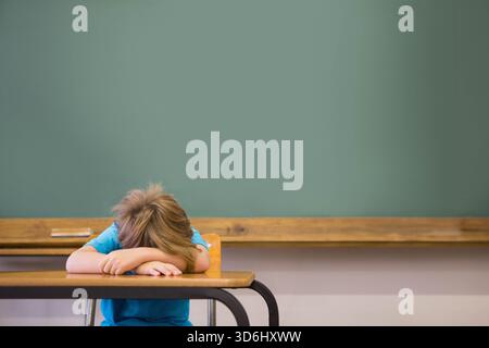 Junge Kind, das Kopf auf den Armen am Schreibtisch im Klassenzimmer mit grüner Tafel und hölzerner Kreidetablett liegt. Schüler, Einzelgänger, Schule, Lernen, Fokus, ruhig, qu Stockfoto