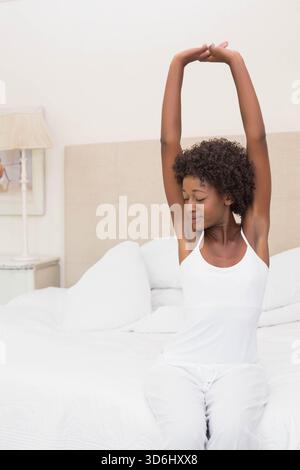 Afroamerikanerin, die auf dem Bett sitzt und die Arme über dem Schlafzimmer spannt, trägt weiße Nachtwäsche. Bettwäsche, Kopfteil, Kissen, Lampe, Kunstwerk, ruhig, ne Stockfoto
