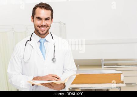 Männlicher Arzt mit Laborkittel, Stethoskop, Bräunungsdiagramm auf der Station mit Bahre, Kopierraum. Arzt, Krankenhaus, Medizin, Dokumentation, Papier Stockfoto