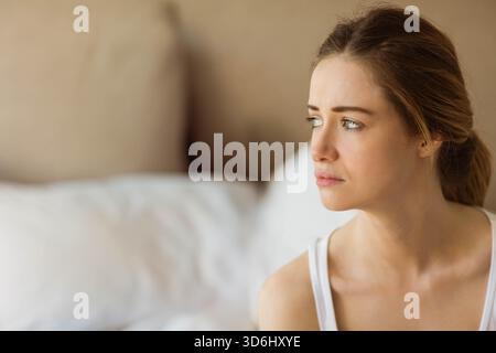 Frau in den späten Zwanzigern, die auf dem Bett sitzt und nach rechts zeigt, mit weißem Tanktop, nachdenklich, Kopierraum. Person, Intim, Porträt, Schlafzimmer, Natur, weich, mit Stockfoto