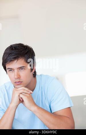 Mann, der sich beugt und Hände unter dem Kinn im Wohnzimmer neben dem Sofa sitzt und ein blaues T-Shirt trägt. Besinnlich, innen, minimalistisch, ruhig, nachdenklich Stockfoto