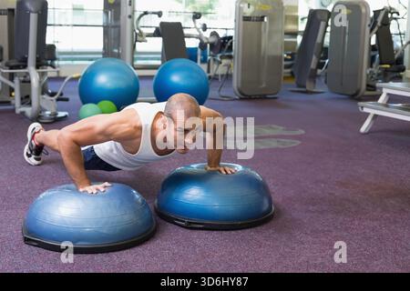 Männlich in den späten 20ern, die Liegestütze auf blauen BOSU-Sportschuhen im Fitnessstudio mit weißem Tanktop machen. Gleichgewicht, Kern, Kraft, funktionell, Fitness, Einrichtung, Betrieb Stockfoto