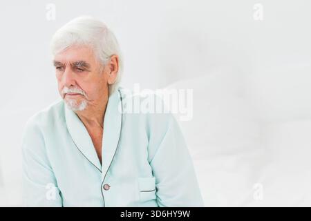 Ein älterer Mann, der im hellen Schlafzimmer auf dem Bett saß, trug einen blauen Bademantel und berührte weiße Kissen. Ältere Menschen, besinnlich, minimalistisch, ruhig, innen, Stockfoto