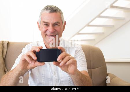 Älterer Mann im Wohnzimmer, der auf dem Sessel sitzt und das Telefon geneigt hält, zeigt Eheringe in der Nähe der Treppe. Ältere Menschen, Smartphone, Sofa, Sonnenlicht, Innenraum, Mini Stockfoto