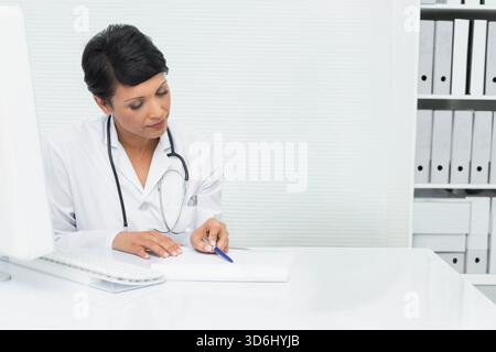 Asiatische Ärztin in weißem Labormantel, die am Schreibtisch sitzt und mit blauem Stift und Stethoskop schreibt. Klinisch, medizinisch, Arbeitsplatz, Notebook, Monitor, o Stockfoto