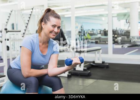 Frau, die im Fitnessstudio auf blauem Stabilitätsball sitzt, sich in der Nähe von Spiegelwänden mit blauer Kurzhantel kräuselt. Fitness, Training, Kraft, Bewegung, Workout, Konzentration, Motivation Stockfoto