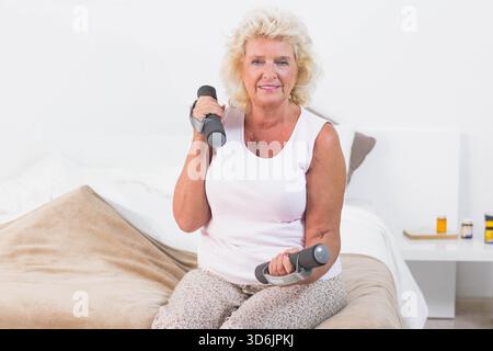 Ältere Frau, die auf dem Bett sitzt und kleine Hanteln in der Nähe von Nachttisch-Medikamenten hegt. Gewichte, sitzend, reif, Schlafzimmer, minimalistisch, Wellness, fi Stockfoto