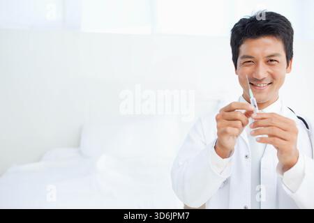Asiatischer Mann im mittleren Erwachsenenalter mit weißem Mantel und Stethoskop, die Spritze in der Nähe des Untersuchungsbettes, Kopierraum. Arzt, Injektion, Klinik, steril, realistisch, reparieren Stockfoto