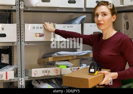 Der Mitarbeiter in rotem Oberteil greift nach dem gekennzeichneten weißen Behälter auf dem Rack, hält die Pappverpackung und den gelben Scanner im Lagerraum mit verschiedenen Artikeln. Stockfoto