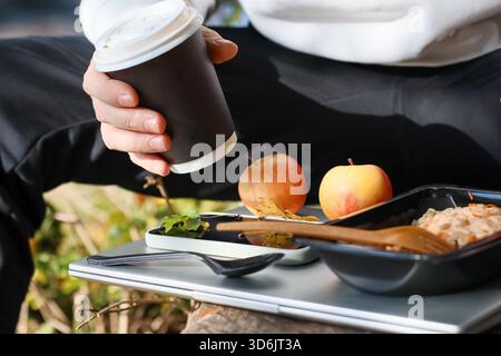 Die Hand der Person greift eine dunkle Kaffeetasse über Äpfeln, Smartphone, Utensilien und Behälter mit gemischtem Reis und Gemüse auf einem Tablett im Park. Stockfoto