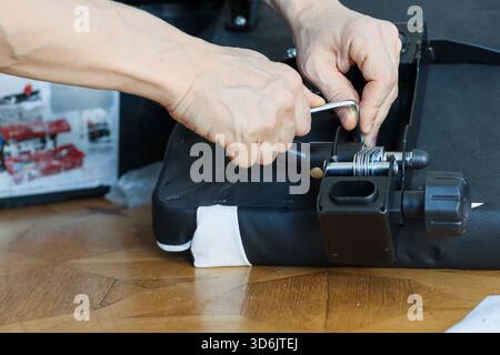 Nahaufnahme der Hände mit einem Schraubenschlüssel zum Einstellen und Festziehen der Federn an einem schwarzen Sitzmechanismus, umgeben von Montageanweisungen auf einem Holzboden. Stockfoto