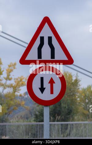 Verkehrsschilder warnen Fahrer vor einer engen Straße und erfordern Priorität für entgegenkommende Fahrzeuge auf einer Zweibahnstraße Stockfoto
