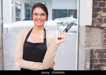 Erwachsene Stylistin mit belüfteter Bürste, schwarzer Schürze, durchsichtiger Brille im Salon. Fenster, Schaufenster, Urban, Backstein, Marmor, modern, mit Flair Stockfoto