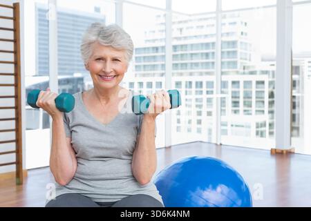 Eine ältere Frau, die im Fitnessstudio türkisfarbene Kurzhanteln hob, ein hellgraues T-Shirt und eine dunkle Leggings trug. Urban, Hochhaus, Skyline, Tageslicht, Fenster, Mini Stockfoto