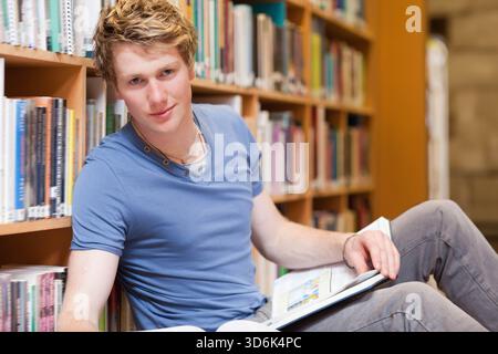 Erwachsener männlicher Student sitzt auf dem Bibliotheksboden und lehnt sich an ein Bücherregal und liest ein offenes Lehrbuch. Mann, Leser, Wirbelsäule, Regale, Studie, warm, glühen Stockfoto
