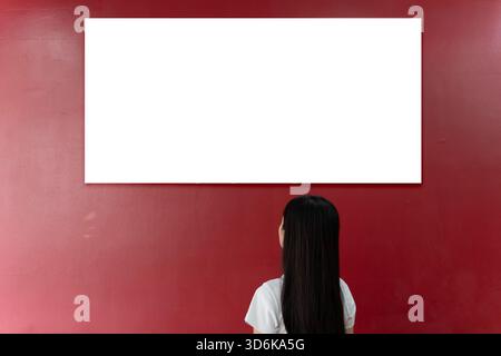 Frau, die vor einem leeren weißen Schild an einer roten Wand steht und einen sauberen Mockup-Raum für Design, Werbung, Branding, Nachrichtenanzeige, mar bietet Stockfoto