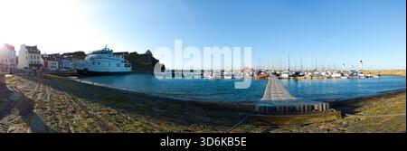 Breite Hafenlandschaft mit einem langen Pier, der sich in ruhiges blaues Wasser erstreckt, gesäumt von verankerten Booten und einem entfernten Leuchtturm. Helle Sonne, Küstenstadt-Atmosphäre Stockfoto
