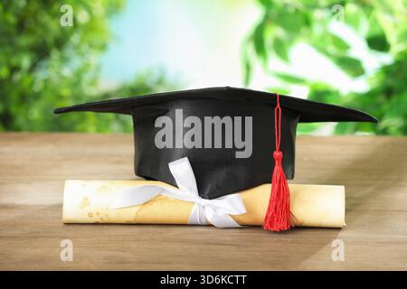 Graduierungskappe und Diplom auf Holztisch vor verschwommenem Hintergrund, Nahaufnahme Stockfoto