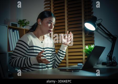 Stressige Frau mit Laptop und schnurlosen Ohrstöpseln, die nachts Videoanrufe tätigen Stockfoto