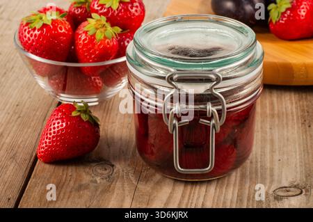 Glasbehälter mit Erdbeermarmelade auf einem Holztisch mit frischen Erdbeeren im Hintergrund, wodurch ein natürliches Obst und hausgemachte Lebensmittel-Konzept ide entsteht Stockfoto