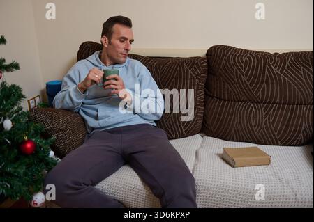 Ein Mann in einem blauen Hoodie sitzt auf einem gemusterten Sofa und schlürft Kaffee, während er an einem Laptop arbeitet. Ein Weihnachtsbaum und eine Geschenkbox sorgen für eine gemütliche Atmosphäre. Stockfoto