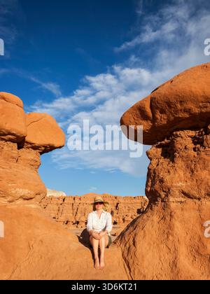Eine Frau mit breiter Krempe und Sonnenbrille sitzt barfuß auf warmem rotem Sandstein zwischen Hoodoo-Felsformationen im Goblin Valley State Park Stockfoto