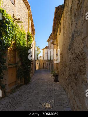 Enge Kopfsteinpflasterstraße im historischen Zentrum von Peratallada, gesäumt von rustikalen Steinhäusern und kletterndem Efeu unter klarem blauen Himmel. Stockfoto