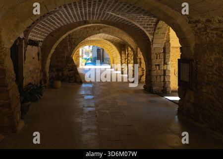 Mittelalterliche Arkade aus Stein an der Plac de les Voltes in Peratallada mit gewölbten Bögen, rustikalen Texturen und sanftem Sonnenlicht, das vom plaza eindringt. Stockfoto