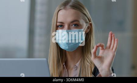 Kaukasische Geschäftsfrau im Gesicht medizinische Maske mit Laptop zeigt OK Zeichen okay Geste Geschäftsfrau Mädchen weibliche Arbeitgeber Manager arbeiten mit Stockfoto