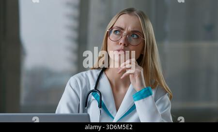 Nachdenkliche kaukasische Frau Doktor denkt, Laptop in Klinik Krankenhaus zu schreiben nachdenklich verwirrte weibliche nachdenkliche Krankenschwester Mädchen denken Idee arbeiten Computer Stockfoto