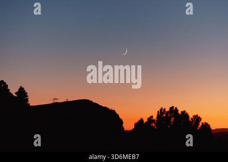 Ein dünner Halbmond über einem leuchtenden orange-blauen Himmel in der Abenddämmerung, silhouettenreiche Bäume und ein Ridgeline im Vordergrund. Friedliche Landschaft im Hintergrund der Dämmerung Stockfoto